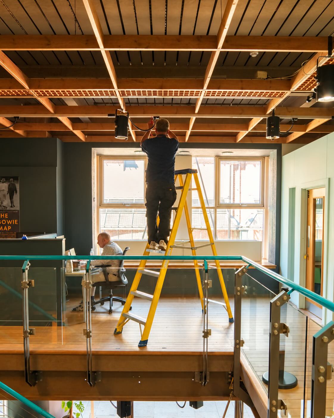 Electrician testing a light fitting on a ladder in an open-plan coworking office